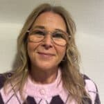 En kvinne med blondt hår og briller smiler til kameraet, iført en rosa og lilla argyle genser, foran en strukturert hvit vegg.
