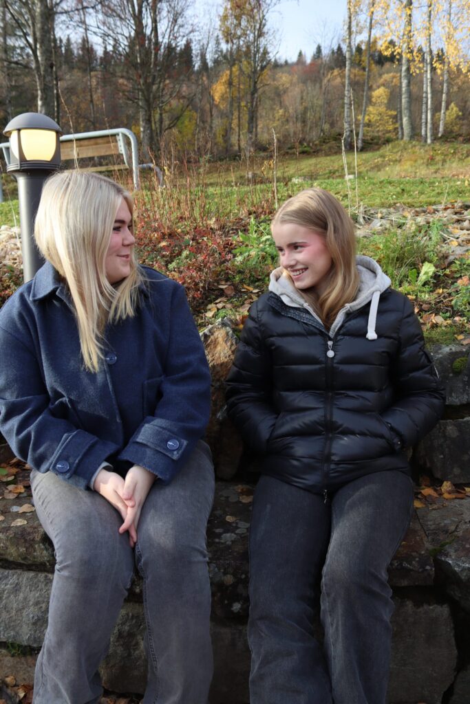 To mennesker sitter på en steinmur utendørs om høsten, vendt mot hverandre og smilende, med trær og løvfall i bakgrunnen.