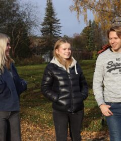 Tre personer står utendørs på en gresslette med høstløv, trær og busker i bakgrunnen, iført uformelle jakker, og snakker sammen.