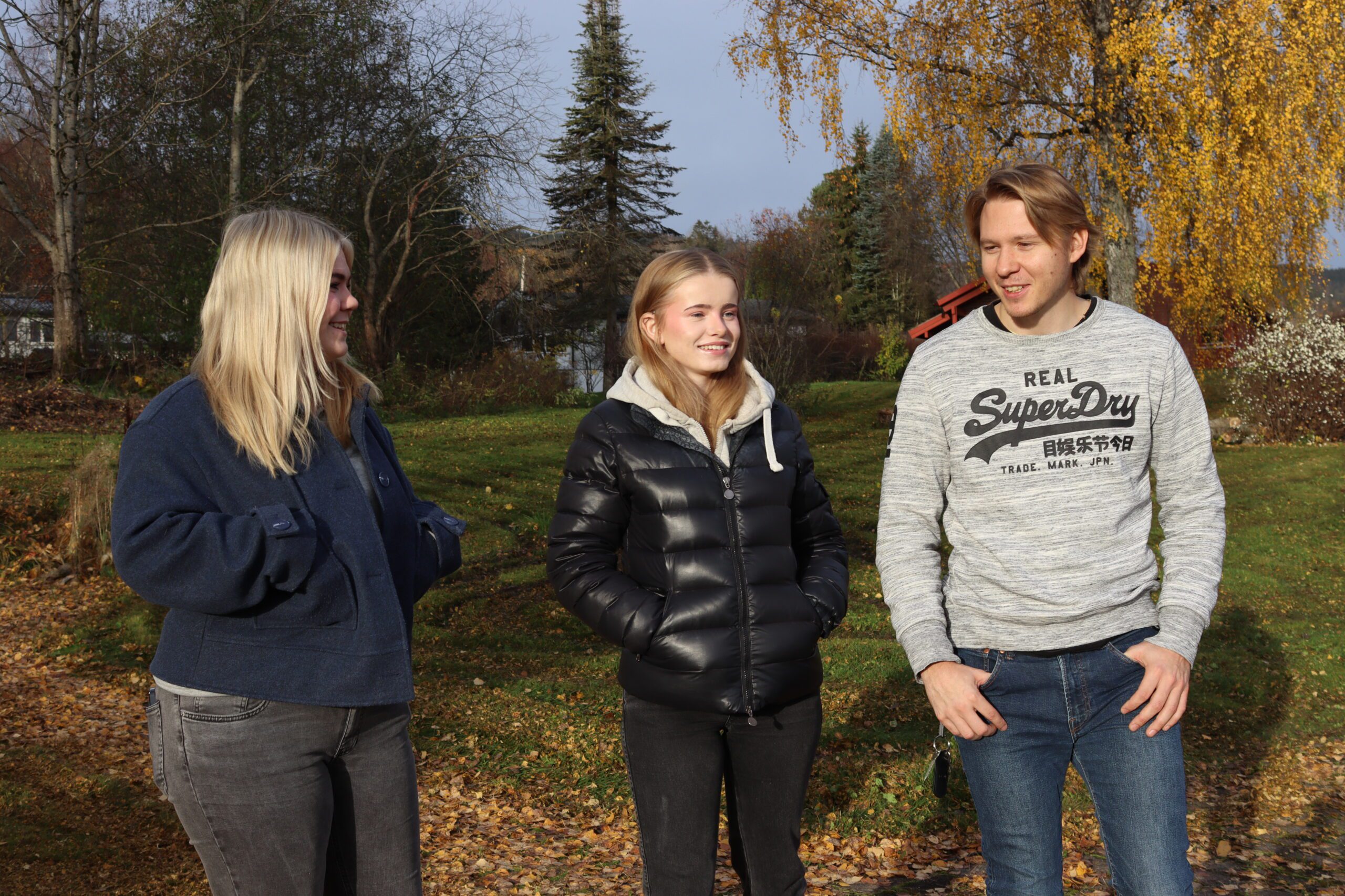 Tre personer står utendørs på en gresslette med høstløv, trær og busker i bakgrunnen, iført uformelle jakker, og snakker sammen.