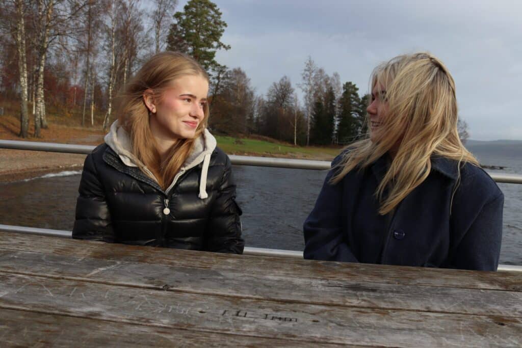 To unge kvinner sitter ved et trebord utendørs ved en innsjø, vendt mot hverandre og smilende, med trær og vann i bakgrunnen.