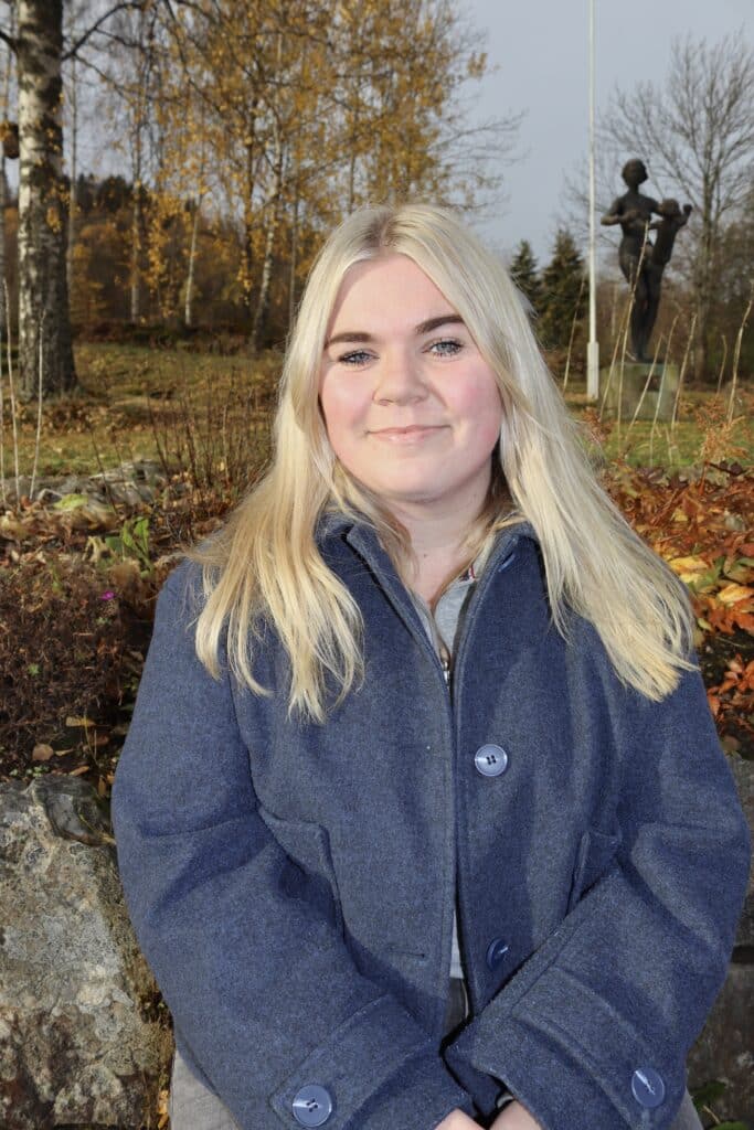 En kvinne med langt blondt hår og en blå frakk smiler ute i en park med høsttrær og en statue i bakgrunnen.
