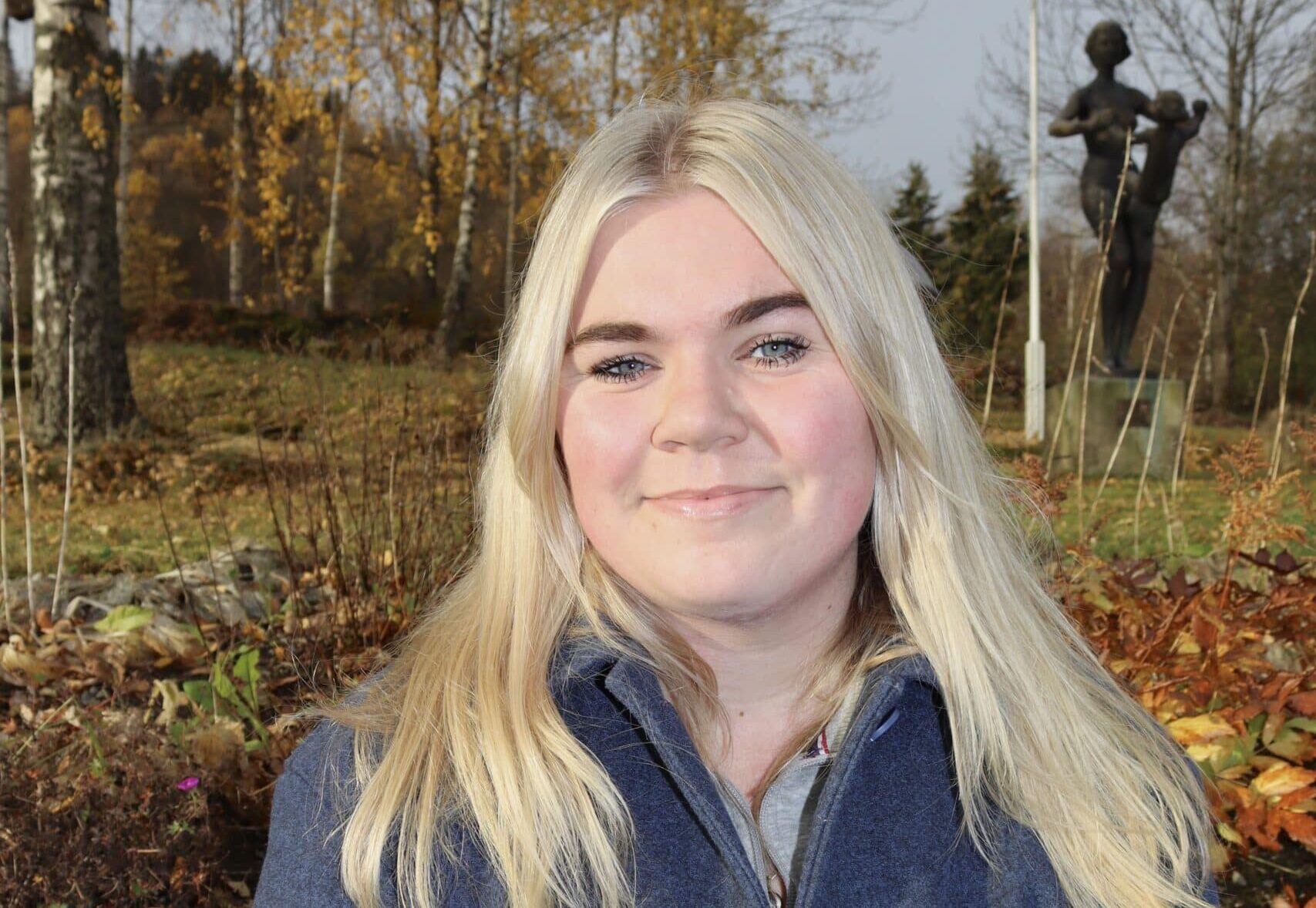 En kvinne med langt blondt hår og en blå frakk smiler ute i en park med høsttrær og en statue i bakgrunnen.