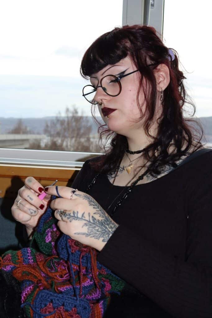 En person med mørkt hår, briller og tatoveringer på hendene sitter innendørs og hekler et fargerikt garnstykke.
