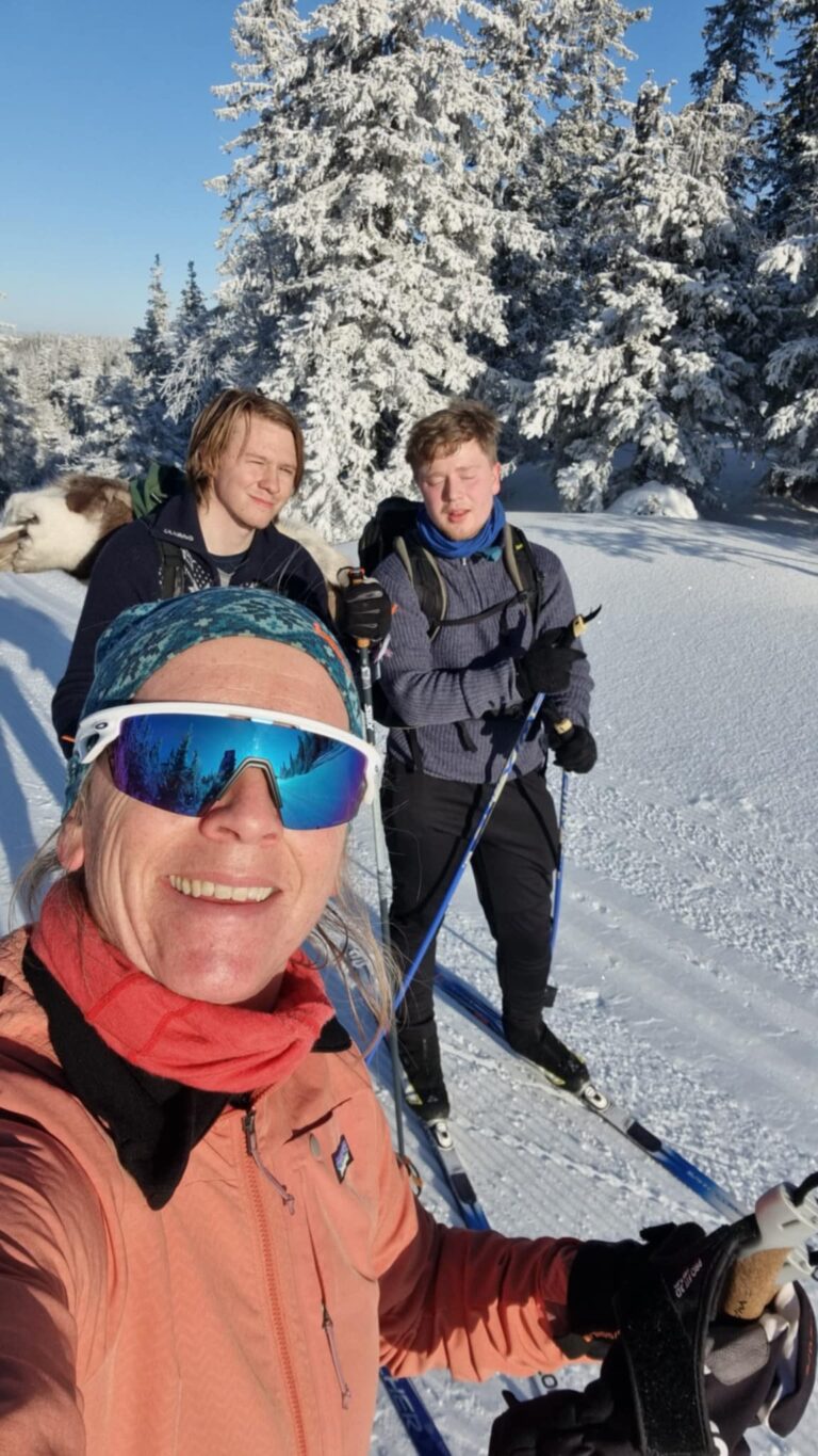 Tre personer poserer for en selfie mens de står på langrennsski i en snødekt løype, omgitt av snødekte trær under en klarblå himmel.