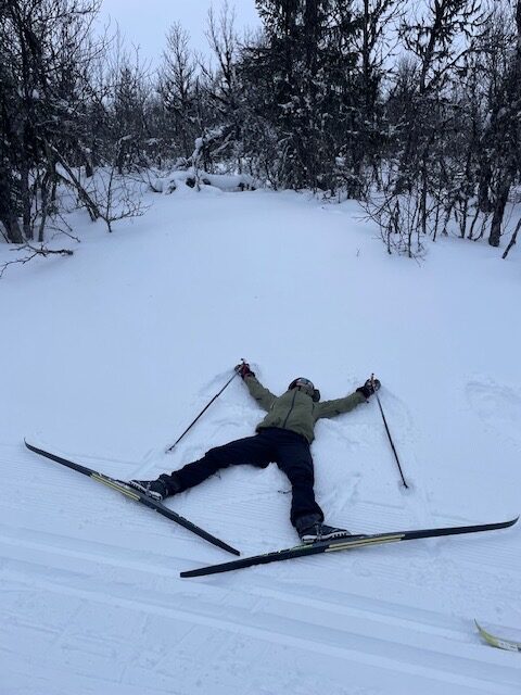 En person i vinterklær ligger på ryggen i snøen med ski og staver spredt ut, omgitt av trær.