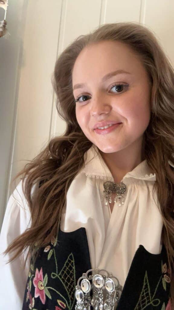 Ung kvinne med langt brunt hår, iført tradisjonell brodert vest, hvit bluse og utsmykkede sølvbrosjer, står foran en vegg med hvitt panel.