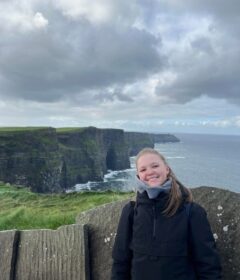 En person i svart jakke står smilende foran Cliffs of Moher, med overskyet himmel og Atlanterhavet i bakgrunnen.