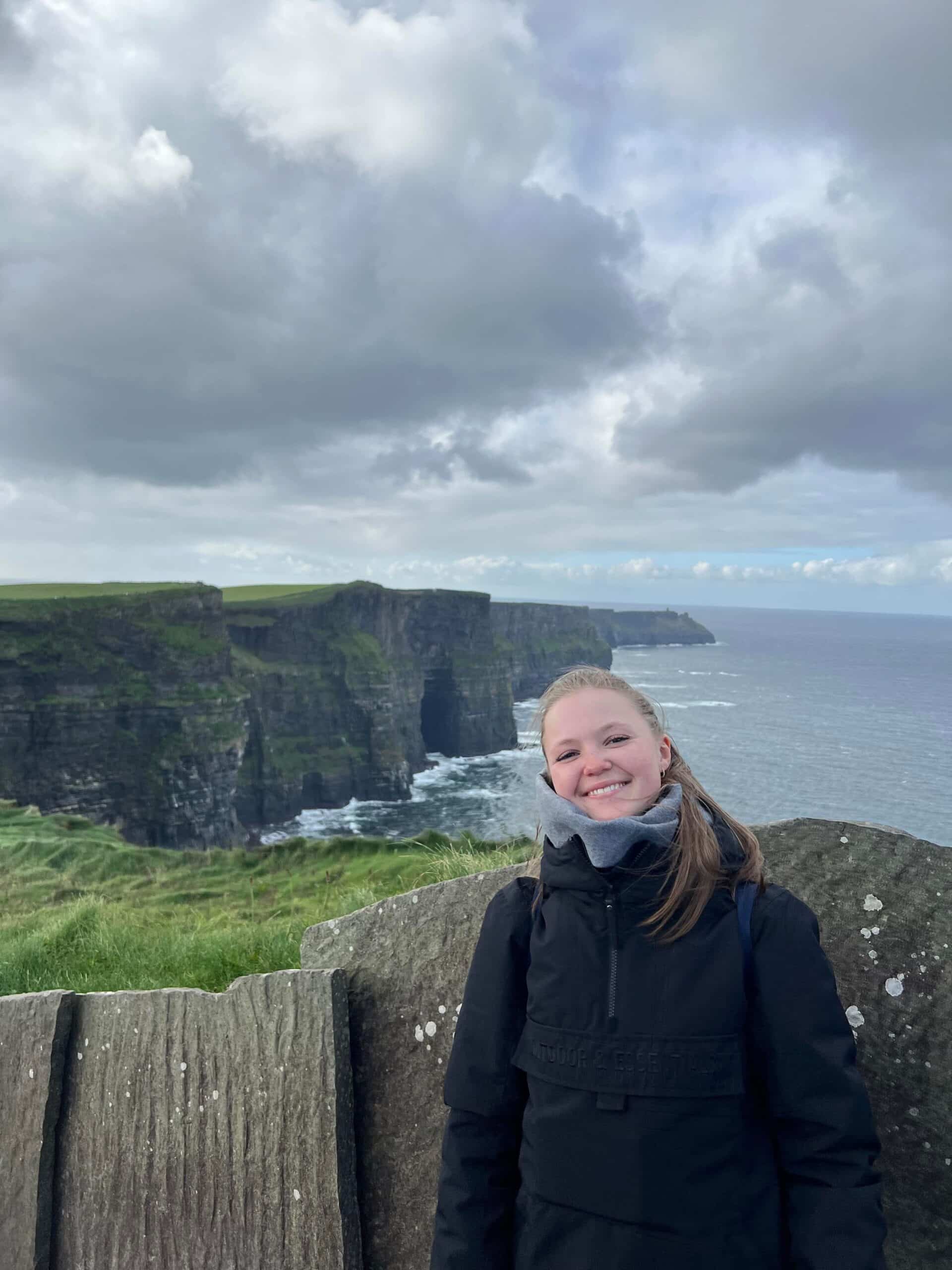 En person i svart jakke står smilende foran Cliffs of Moher, med overskyet himmel og Atlanterhavet i bakgrunnen.