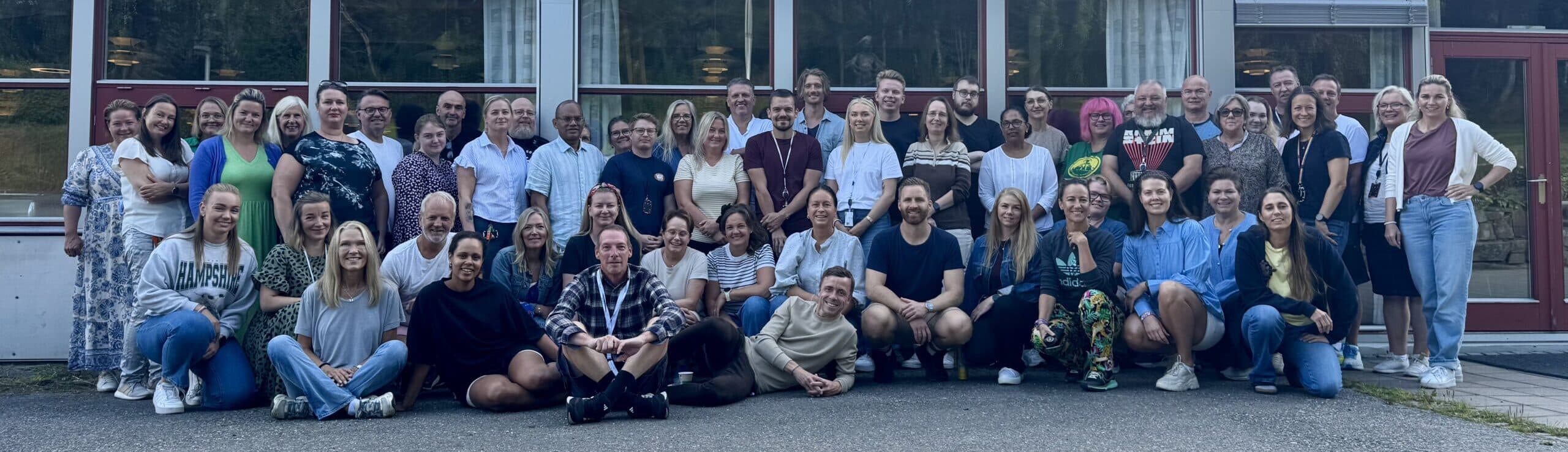 En stor gruppe mennesker, både sittende og stående, poserer sammen utenfor en bygning med store vinduer.