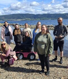 En gruppe på elleve personer og en hund er samlet på en steinete innsjøbredd med trær, vann og åser i bakgrunnen under en delvis overskyet himmel.