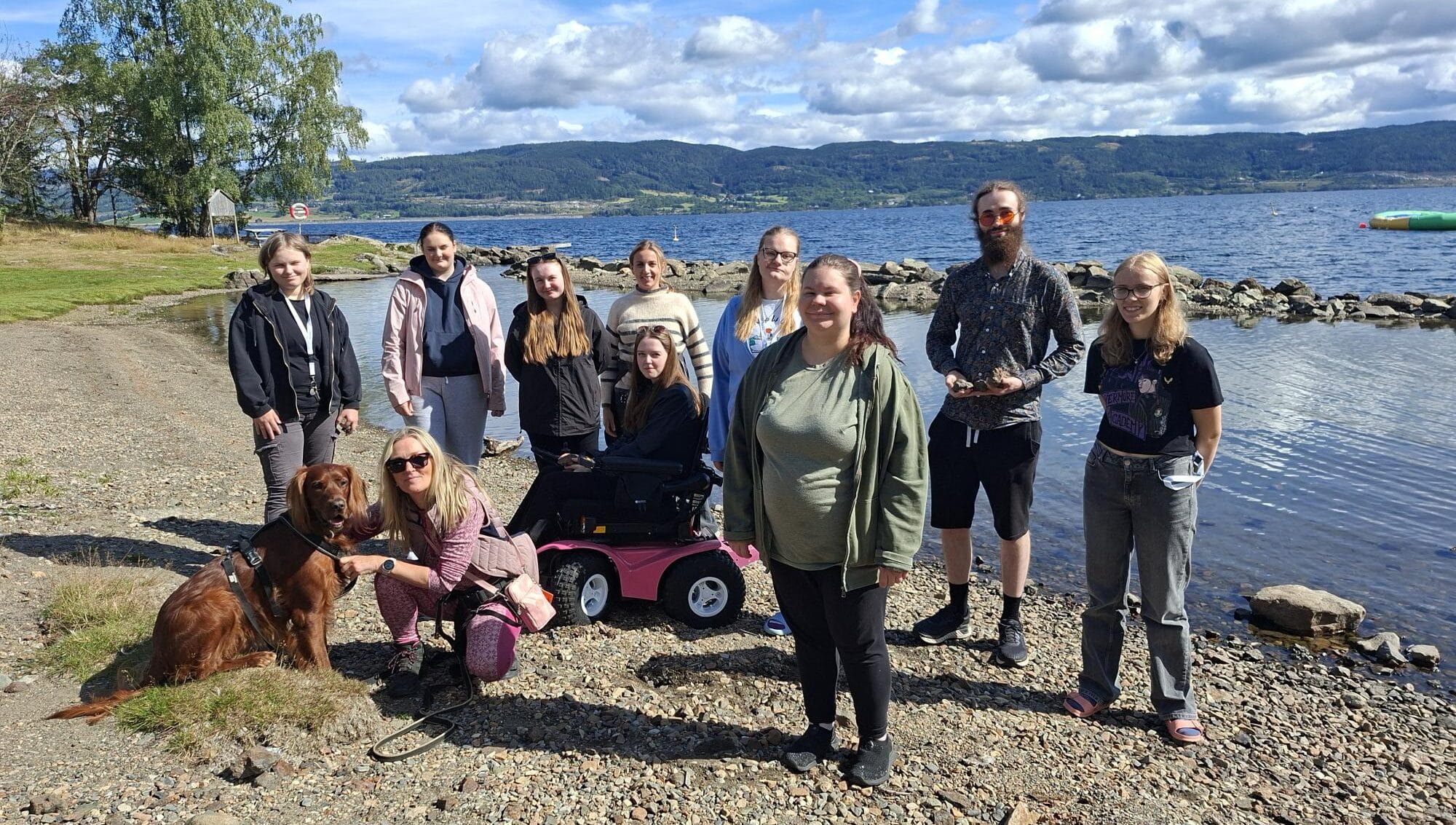 En gruppe på elleve personer og en hund er samlet på en steinete innsjøbredd med trær, vann og åser i bakgrunnen under en delvis overskyet himmel.