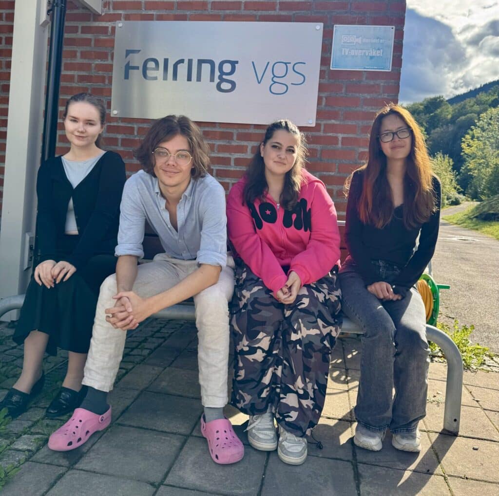20250917_115300466_iOS-1-scaled-e1758185361716 Fire ungdommer sitter på en benk utenfor en murbygning med et "Feiring vgs"-skilt og poserer for et gruppebilde en solfylt dag.