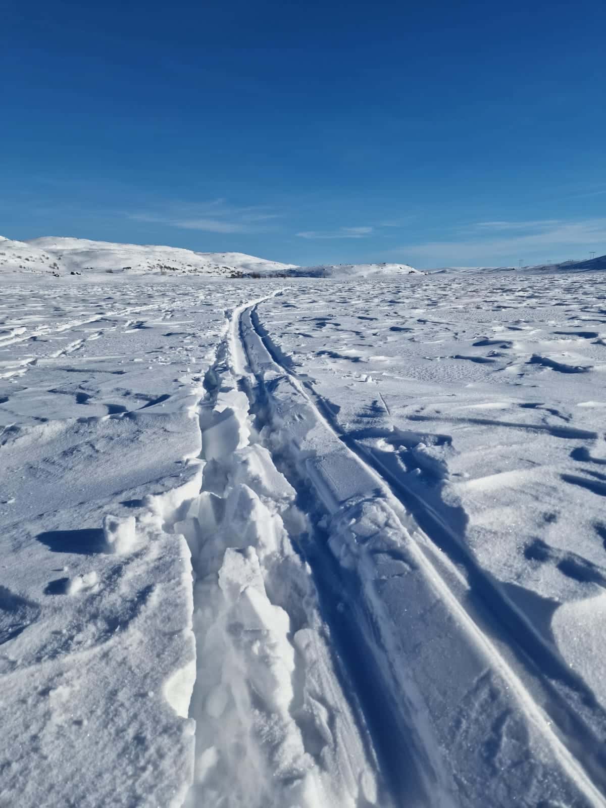 Skispor skjærer seg gjennom et snødekt landskap under en klarblå himmel, med snødekte åser i bakgrunnen.