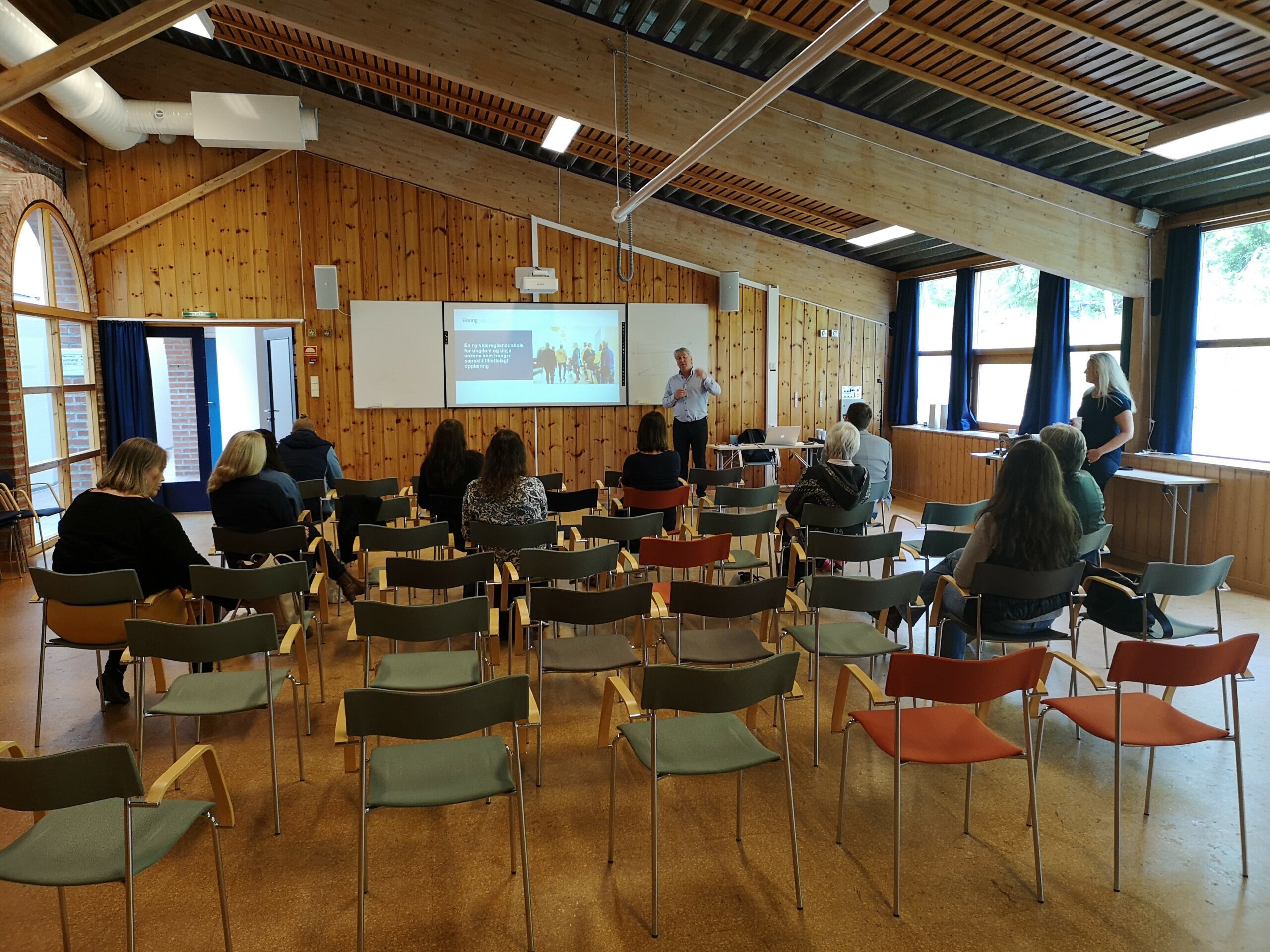 En mann holder en presentasjon for et lite publikum i et rom med trepanel, tomme stolerader og et projisert lysbilde på veggen.