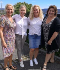 Fire kvinner står sammen ute en solskinnsdag og smiler til kameraet, med trær og åser i bakgrunnen.