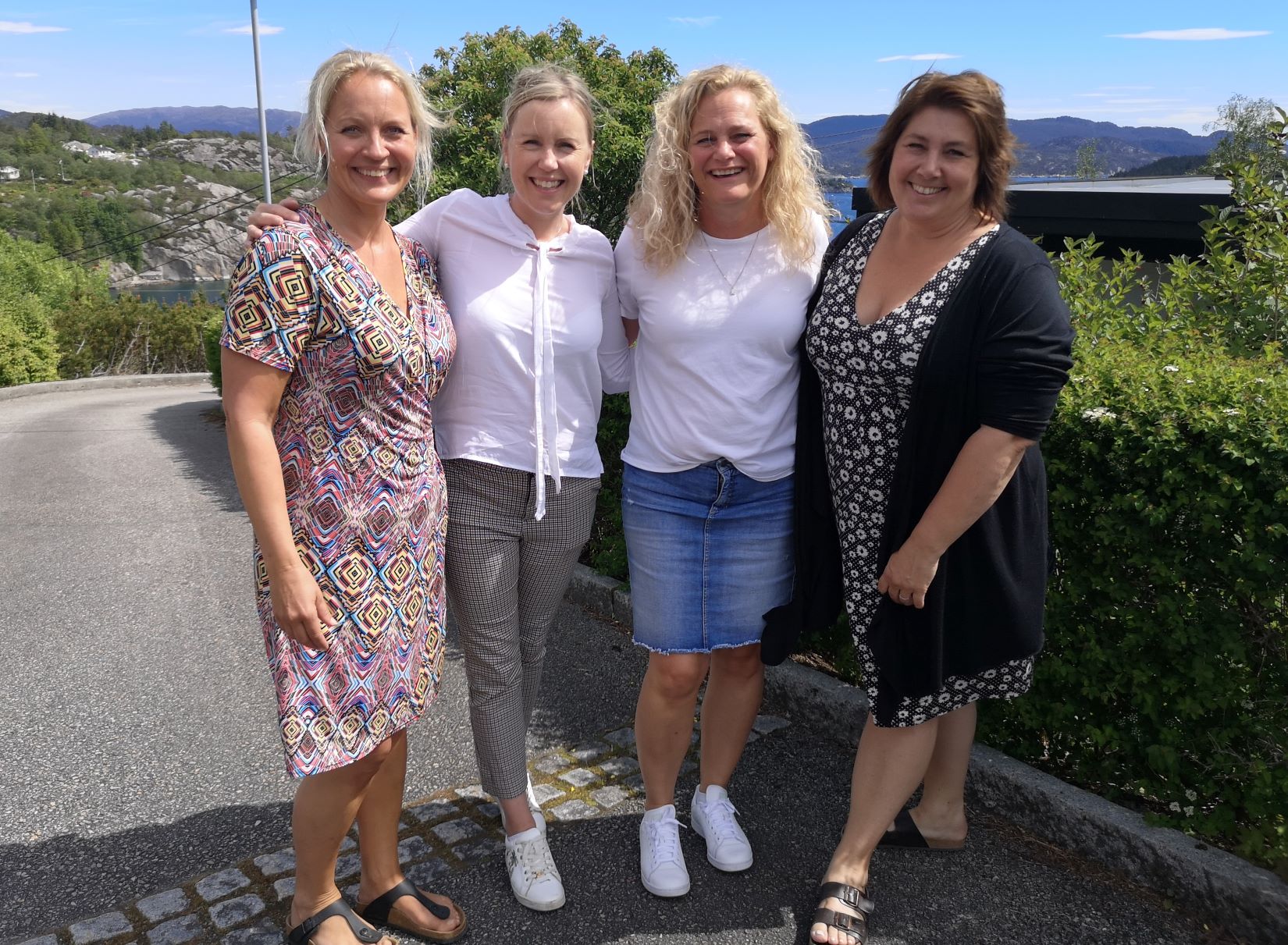 Fire kvinner står sammen ute en solskinnsdag og smiler til kameraet, med trær og åser i bakgrunnen.