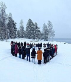En gruppe mennesker står utendørs i snøen og danner et hjerte. Snødekte trær og en vannflate er synlige i bakgrunnen.