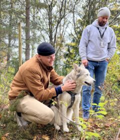 To menn utendørs med en grå og hvit hund; den ene mannen sitter på huk og klapper hunden mens den andre står i nærheten. Trær og løvverk er i bakgrunnen.