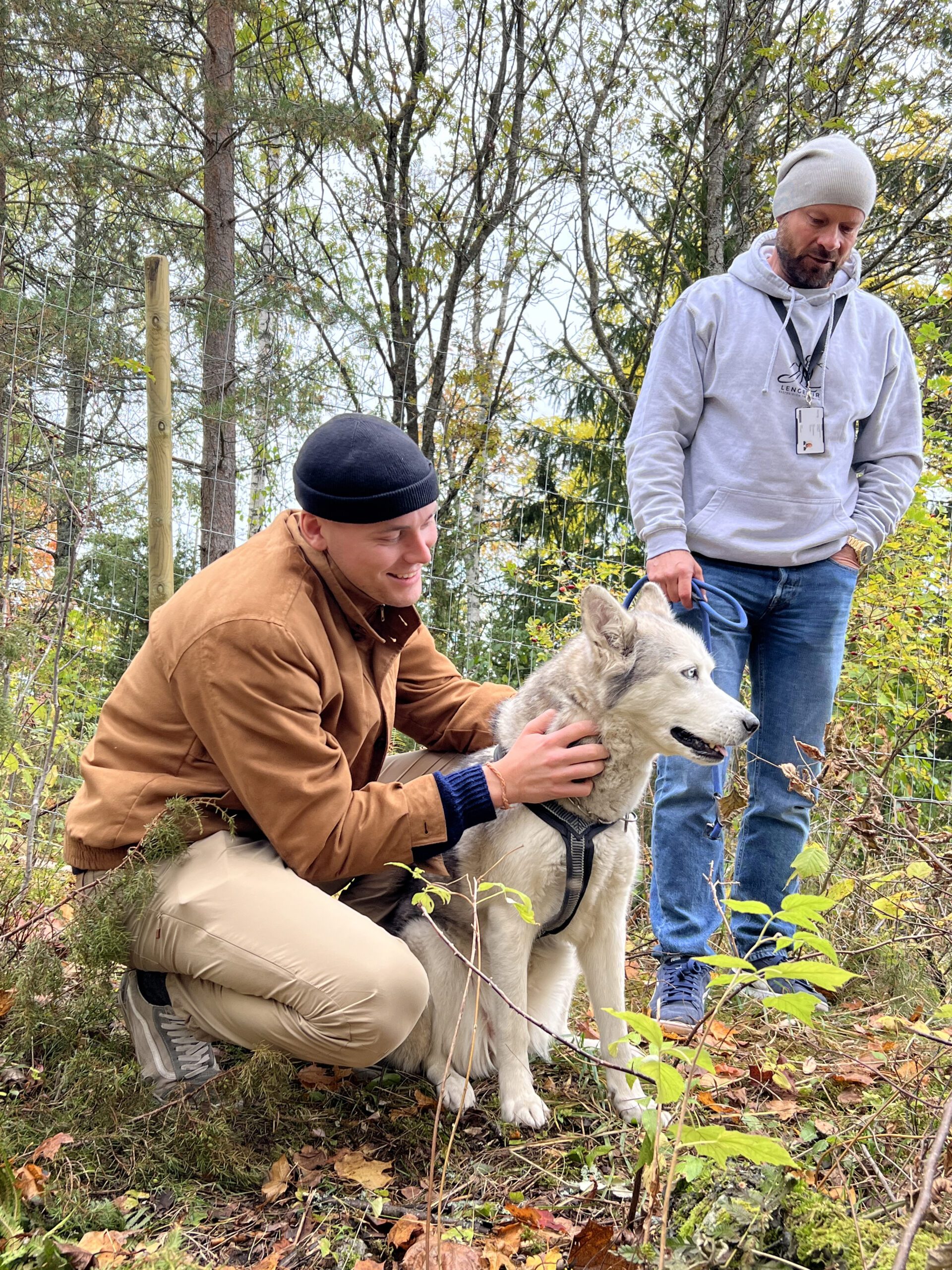 To menn utendørs med en grå og hvit hund; den ene mannen sitter på huk og klapper hunden mens den andre står i nærheten. Trær og løvverk er i bakgrunnen.