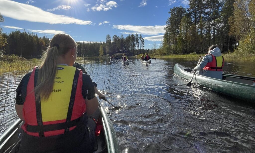 Image-1-scaled Flere personer i redningsvester padler kano på en rolig elv omgitt av trær under en delvis overskyet himmel.