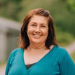 En kvinne med mellomlangt brunt hår smiler utendørs, iført en blågrønn topp. Bakgrunnen er uskarpt grøntområde og en sti.