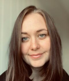 En kvinne med rett, brunt hår og blå øyne smiler mykt til kameraet, med en lys bakgrunn bak seg.