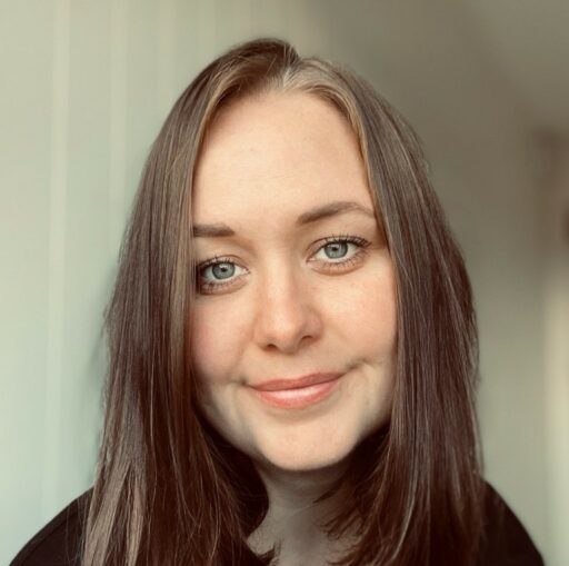 En kvinne med rett, brunt hår og blå øyne smiler mykt til kameraet, med en lys bakgrunn bak seg.