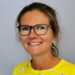 Kvinne med briller og brunt, bakoverstrukket hår, iført gul topp med hvitt blomstermønster, smiler til kameraet mot en lys bakgrunn.