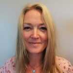 En kvinne med langt blondt hår, iført en lyserosa topp og et halskjede, smiler lett foran en ensfarget bakgrunn.