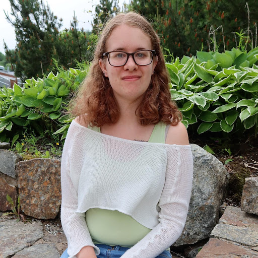 En person med briller og skulderlangt hår sitter utendørs på en steintrapp, omgitt av grønne planter, iført en lysegrønn topp og hvit genser.