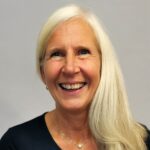 Smilende eldre kvinne med langt hvitt hår, iført mørk topp, fotografert mot en lys bakgrunn.