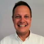 En mann med kort, brunt hår smiler mot kameraet. Han har på seg en hvit skjorte med krage og står foran en ensfarget, lys bakgrunn.