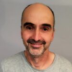 En middelaldrende mann med vikende hårfeste, kort, mørkt hår og trimmet skjegg smiler mot kameraet. Han har på seg en lysegrå skjorte mot en ensfarget bakgrunn.