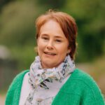 En kvinne med kort, rødlig hår iført grønn genser og mønstret skjerf står utendørs med uskarpt grønt i bakgrunnen.