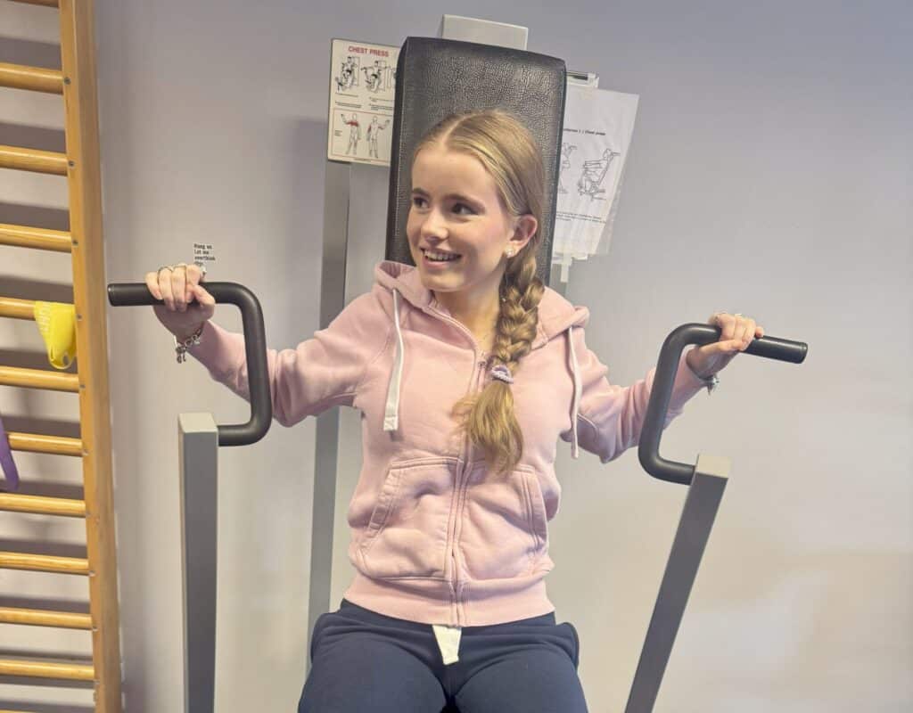 feiring En ung kvinne i rosa hettegenser bruker en brystpressmaskin på et treningssenter, mens hun smiler og ser til siden. På veggen bak henne kan man se instruksjonsskjemaer.