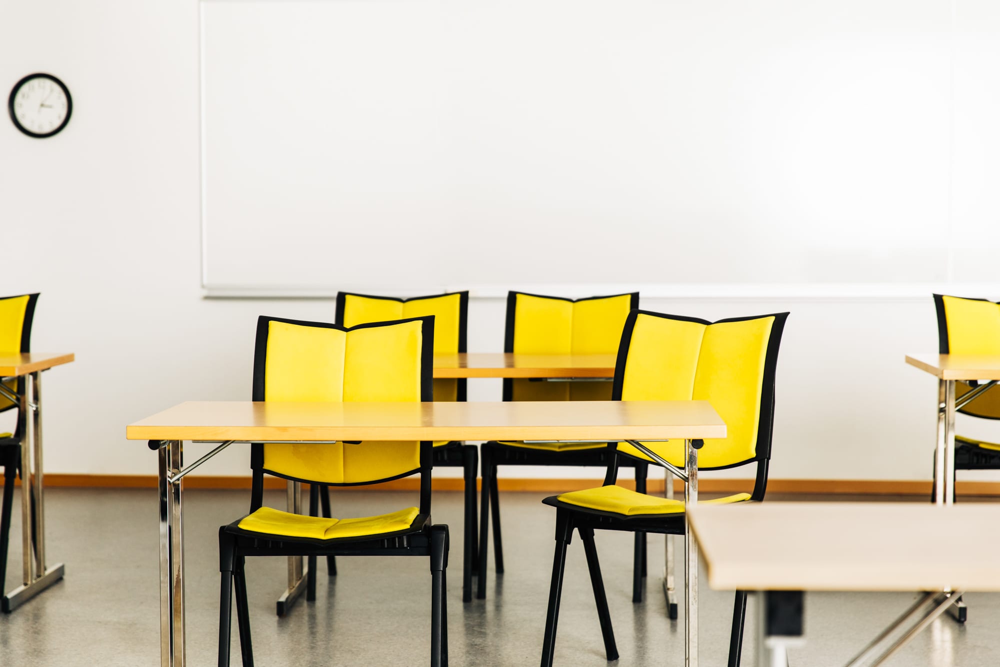 Klasserom med gule stoler og trepulter på rekke og rad, tavle og klokke på veggen i bakgrunnen.