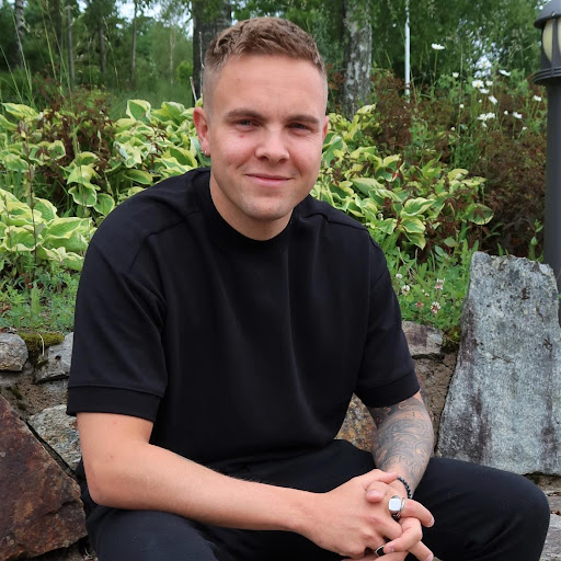 En person med kort hår og svart t-skjorte sitter utendørs foran en steinmur og grønne planter og smiler til kameraet.