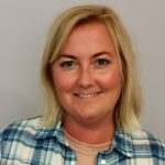 En kvinne med blondt hår smiler til kameraet, iført en blå og hvit rutete skjorte mot en vanlig lys bakgrunn.