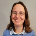 En kvinne med langt brunt hår og briller smiler mot kameraet. Hun har på seg en lyseblå genser over en skjorte med krage, og bakgrunnen er ensfarget lys.