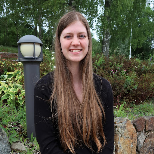 En ung kvinne med langt brunt hår, iført svart topp, sitter utendørs på en steinkant med grønne planter og en lyktestolpe i bakgrunnen og smiler til kameraet.