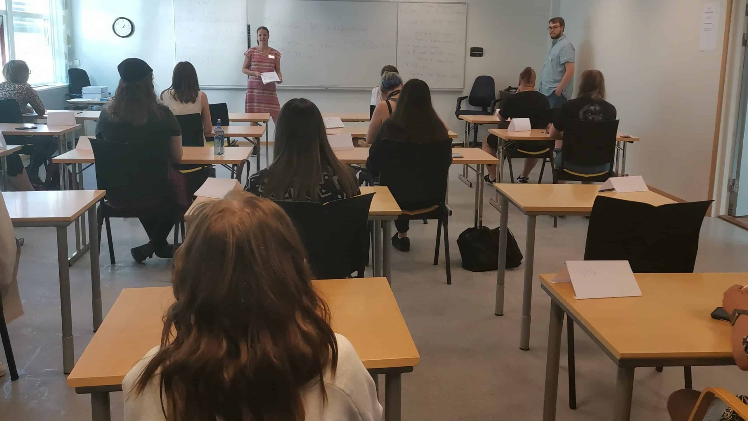 Elevene sitter ved pulter i et klasserom vendt mot en tavle, mens en kvinne står foran og snakker, og en mann står like ved.