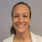 En kvinne med bakoverbøyd, lysebrunt hår og en lys genser smiler til kameraet mot en grå bakgrunn.