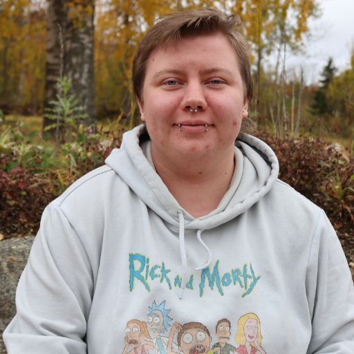 En person med kort hår og piercinger i ansiktet sitter utendørs iført en lysegrå Rick and Morty-hettegenser, med høsttrær og løvverk i bakgrunnen.