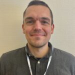 En mann med kort hår og et lite smil har på seg en mørk skjorte med knapper og et nøkkelbånd, og står foran en lys vegg.