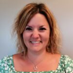 Kvinne med skulderlangt, blondt, bølget hår, smilende, iført grønnmønstret topp og et halskjede med