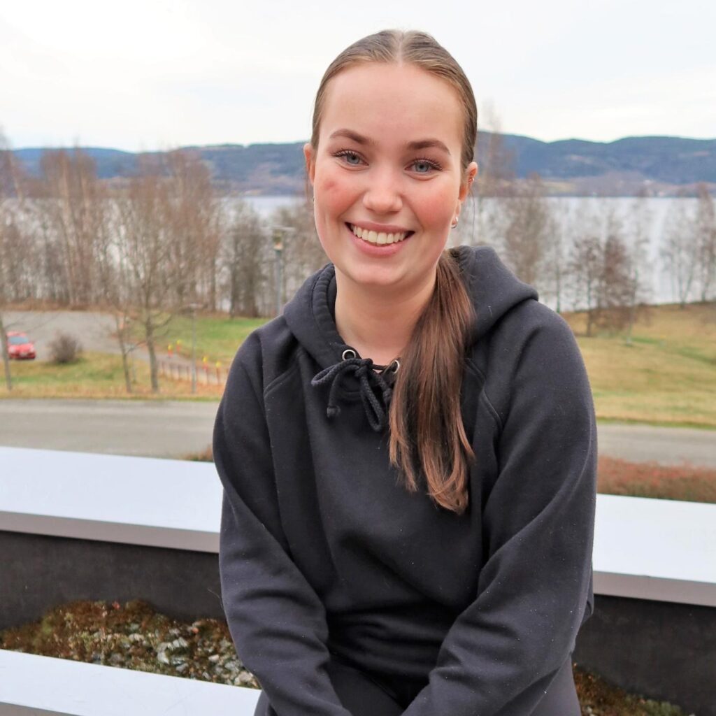 En ung kvinne med hestehale smiler mens hun sitter utendørs foran en innsjø og trær, iført svart hettegenser.