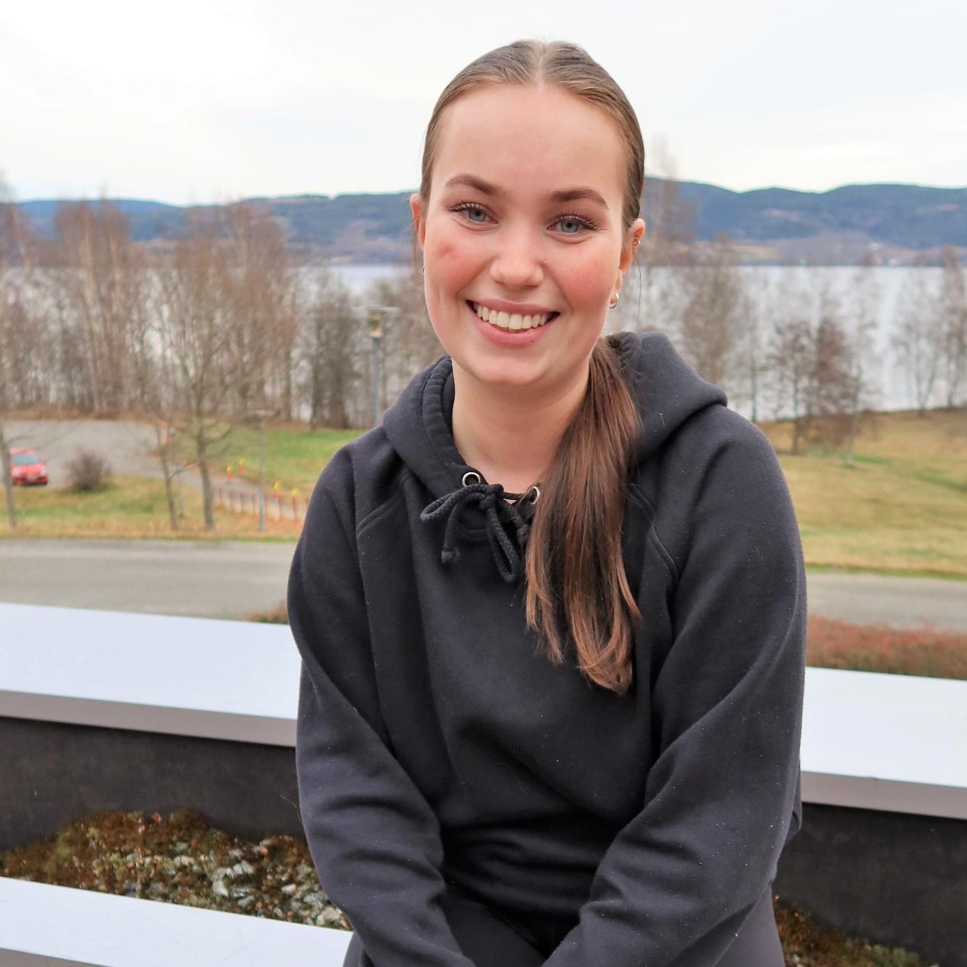 En ung kvinne med hestehale smiler mens hun sitter utendørs foran en innsjø og trær, iført svart hettegenser.