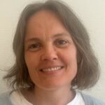 En kvinne med skulderlangt, brunt hår smiler til kameraet mot en vanlig, lys bakgrunn.