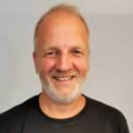 En middelaldrende mann med kort, lyst hår og skjegg smiler mot kameraet. Han har på seg en svart t-skjorte og står mot en ensfarget, lys bakgrunn.